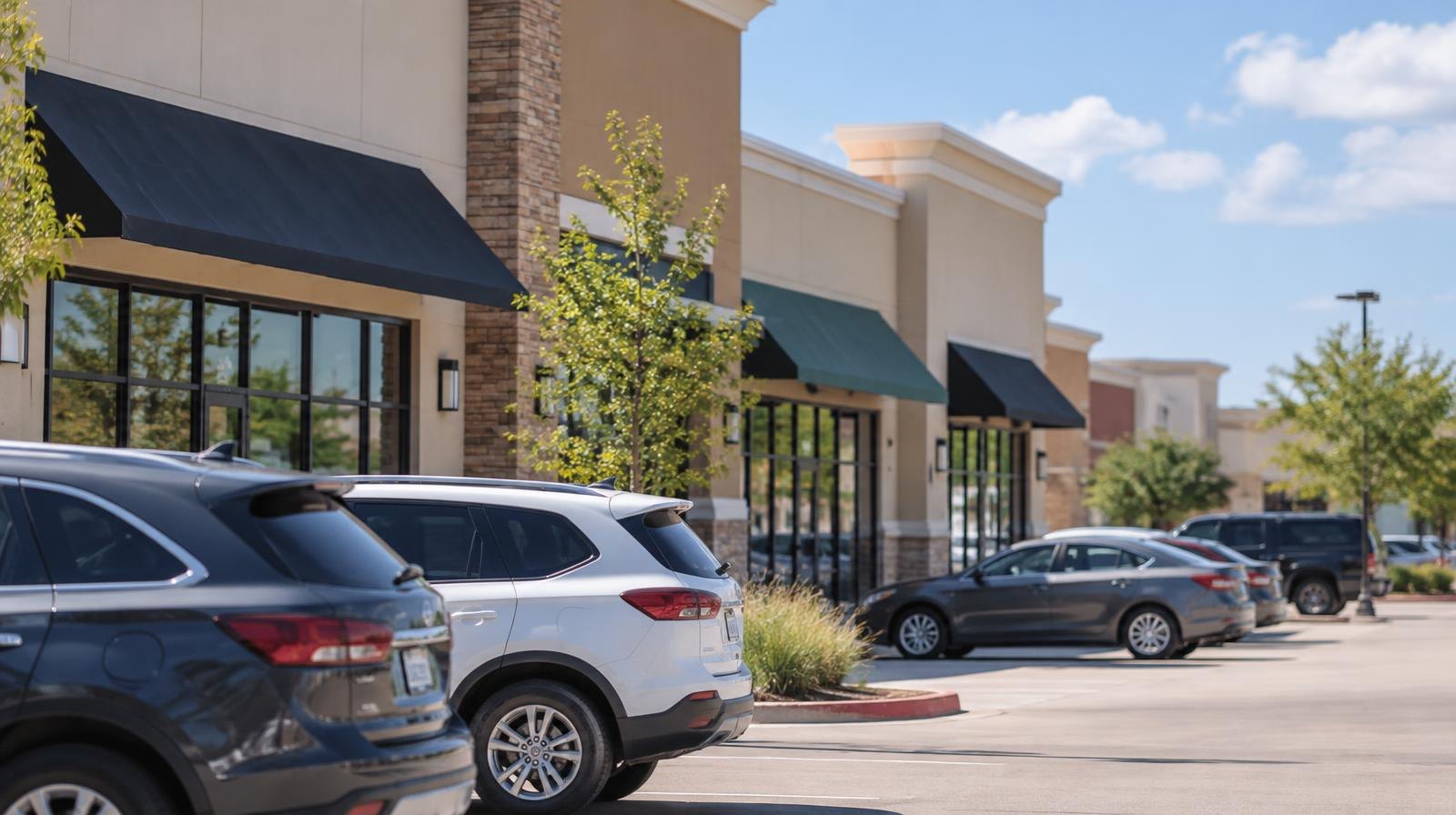 Row of retail storefronts in a suburban shopping center with a large parking lot.