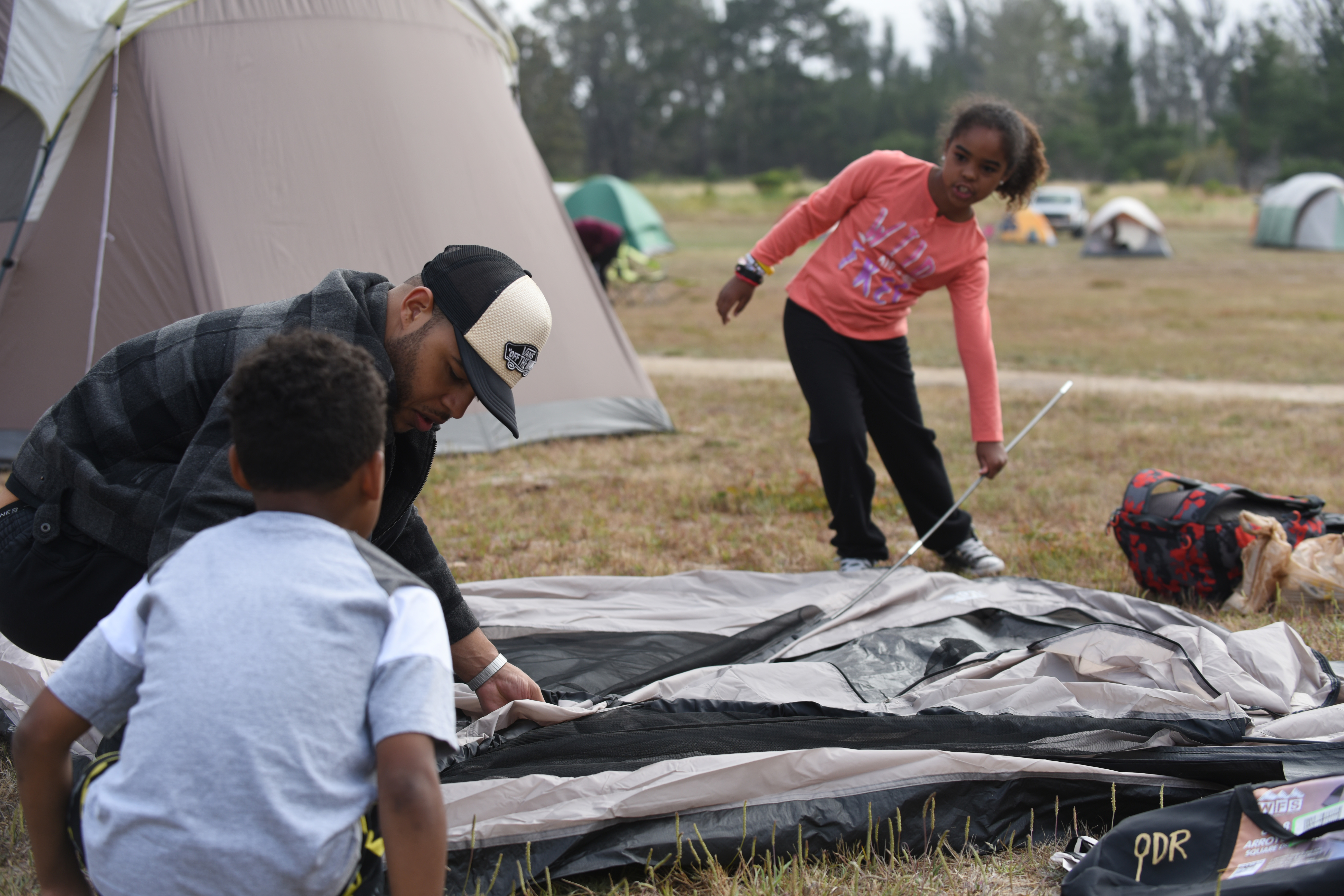 An adult and two children assemble a tent on a grassy field at an outdoor campout.