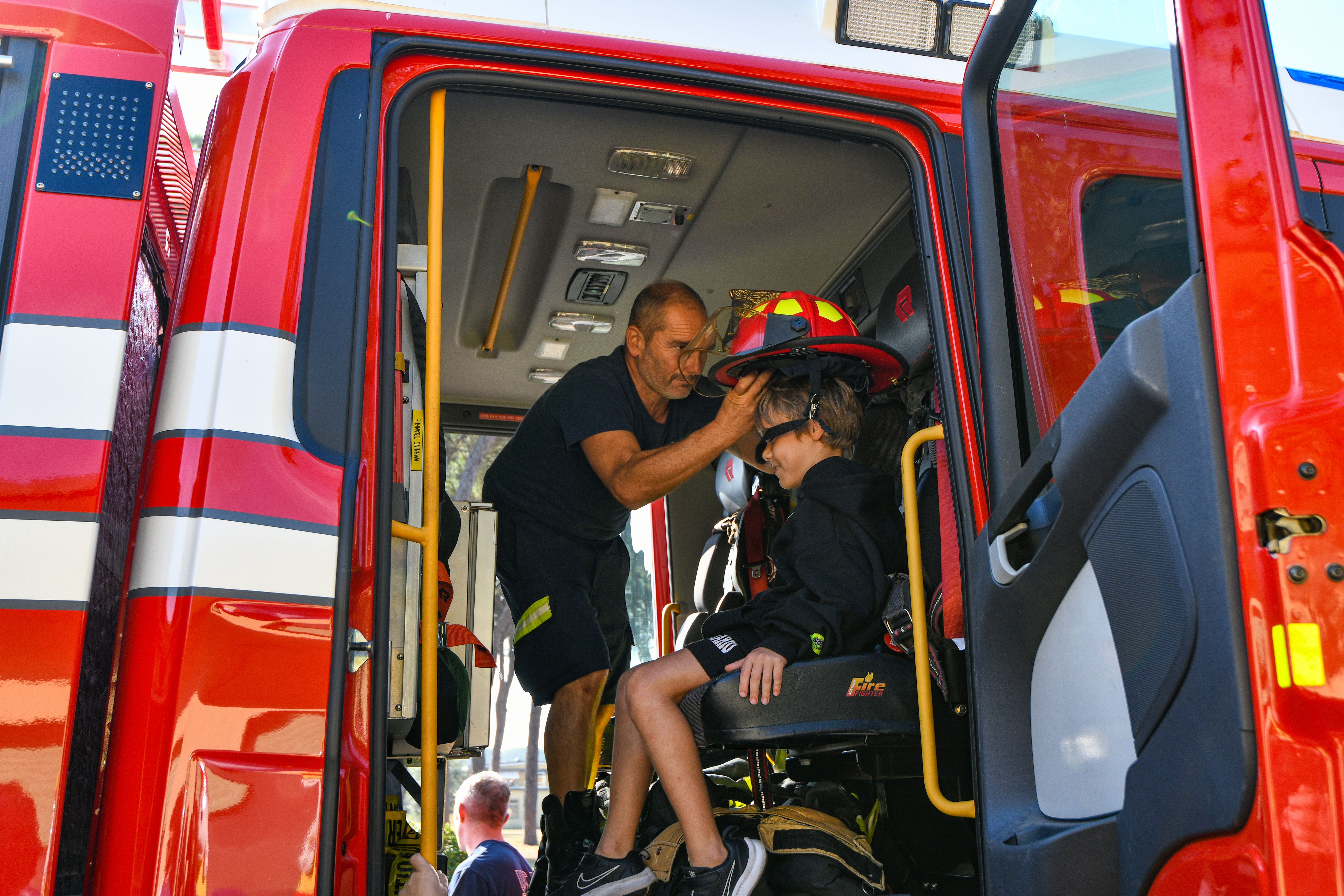 A firefighter places a red fire helmet on a child seated inside a fire truck cab.