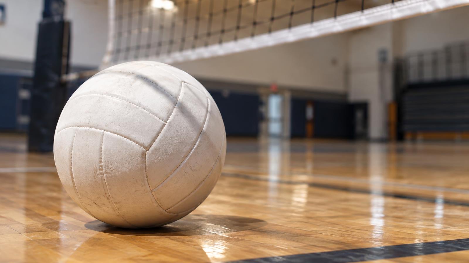 A white volleyball resting on a hardwood gym floor near a net.