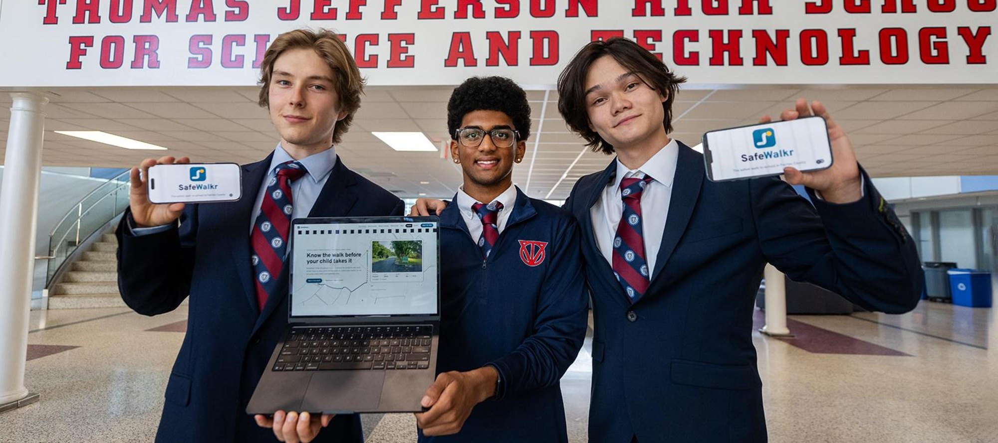 Three teenage students in school blazers hold smartphones and a laptop in a school hallway.