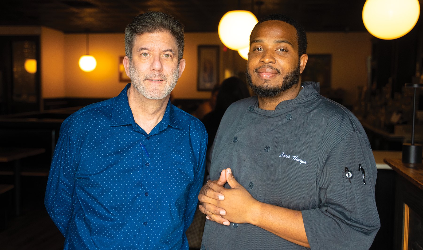 Two men standing together inside a restaurant, one in a blue shirt and one in a dark chef's coat.