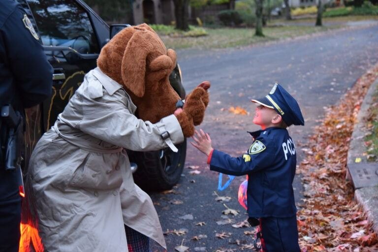 Child in Halloween costume high-fiving a large dog mascot on a leaf-covered street with a police vehicle behind them.