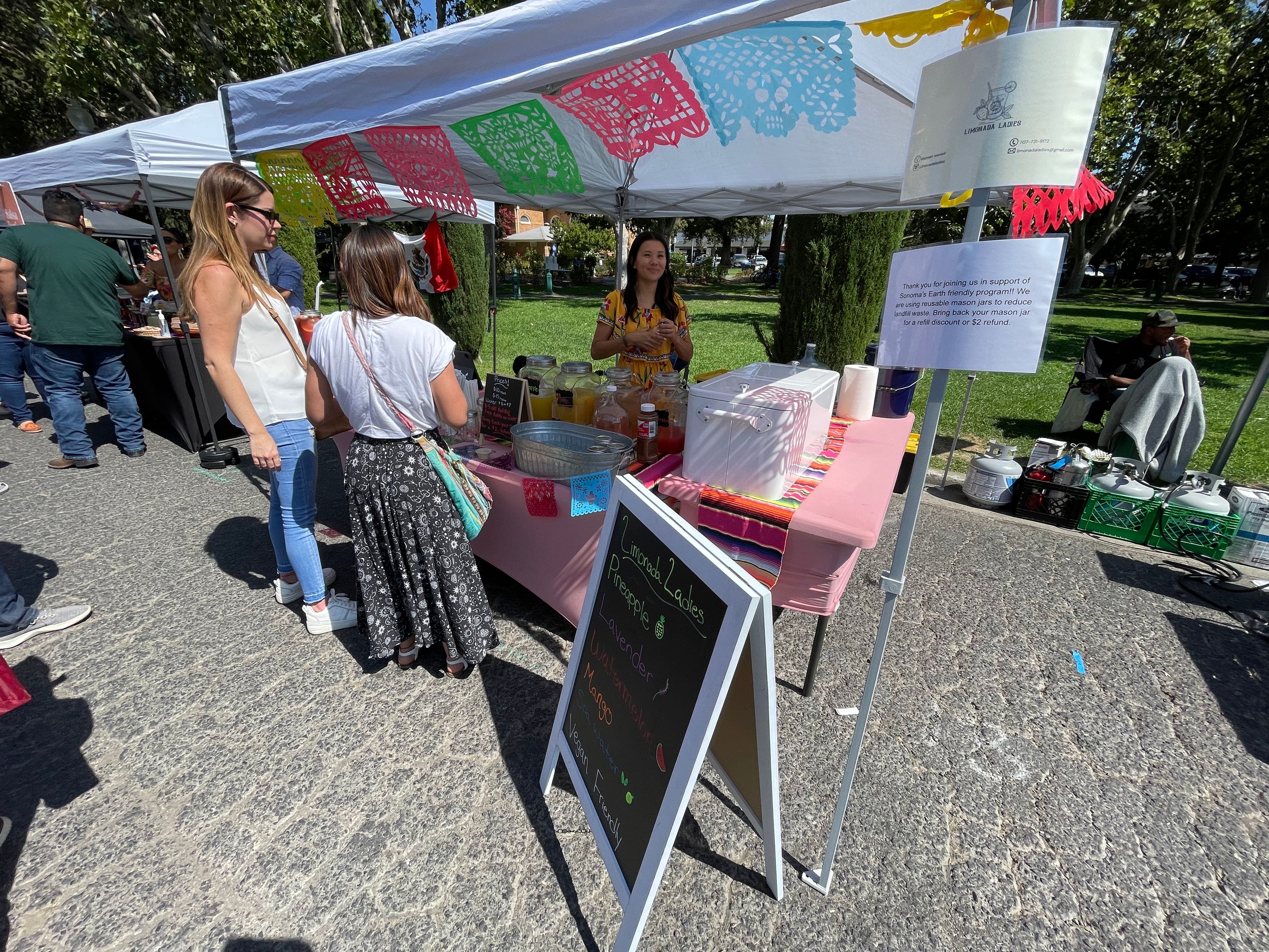 Colorful papel picado banners hang over an outdoor festival booth on a sunny day.