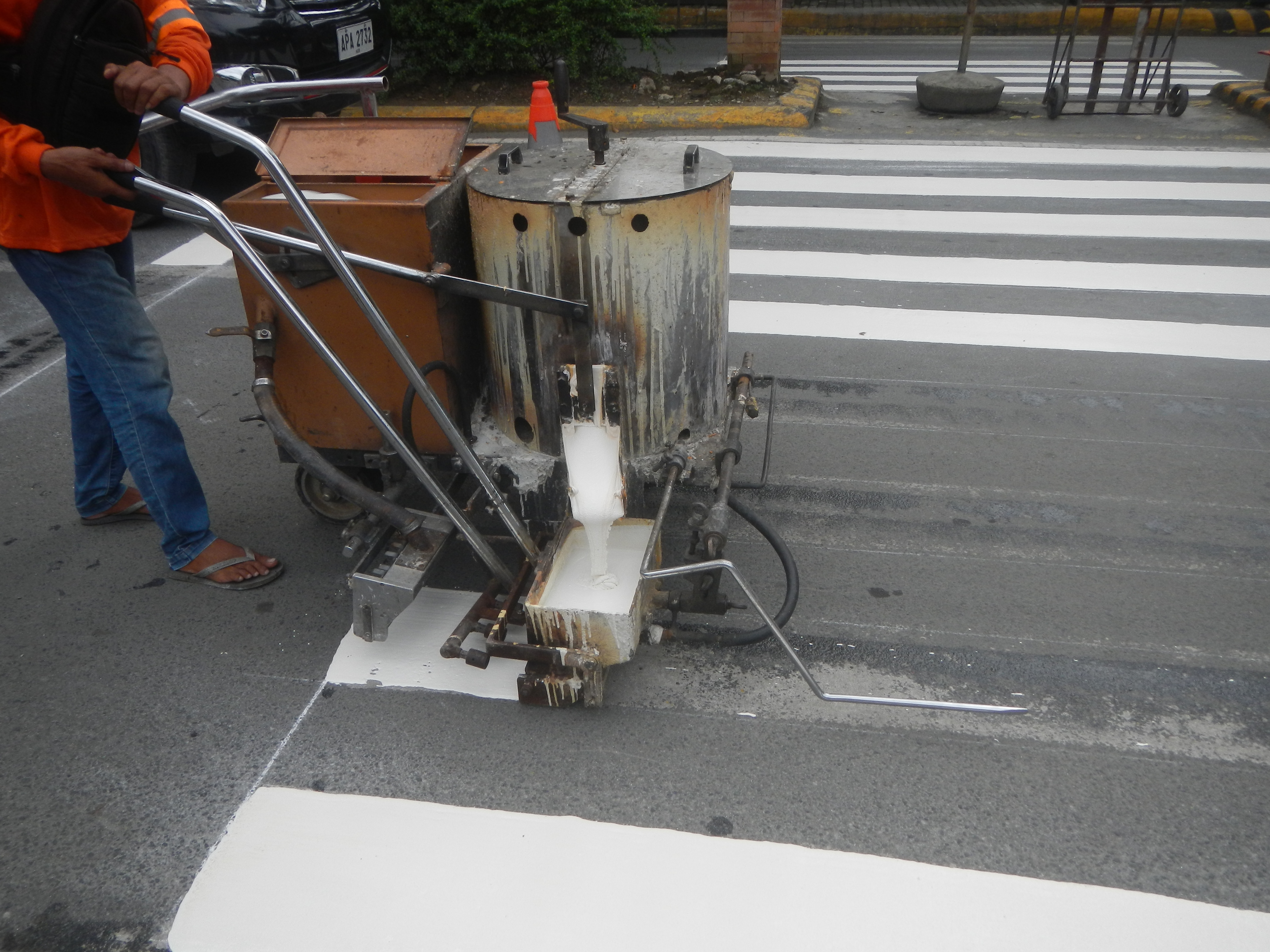 Worker in orange safety vest painting white crosswalk stripes on a road.