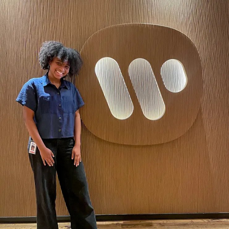 A smiling young woman in a navy shirt stands before an illuminated Warner Music Group logo.