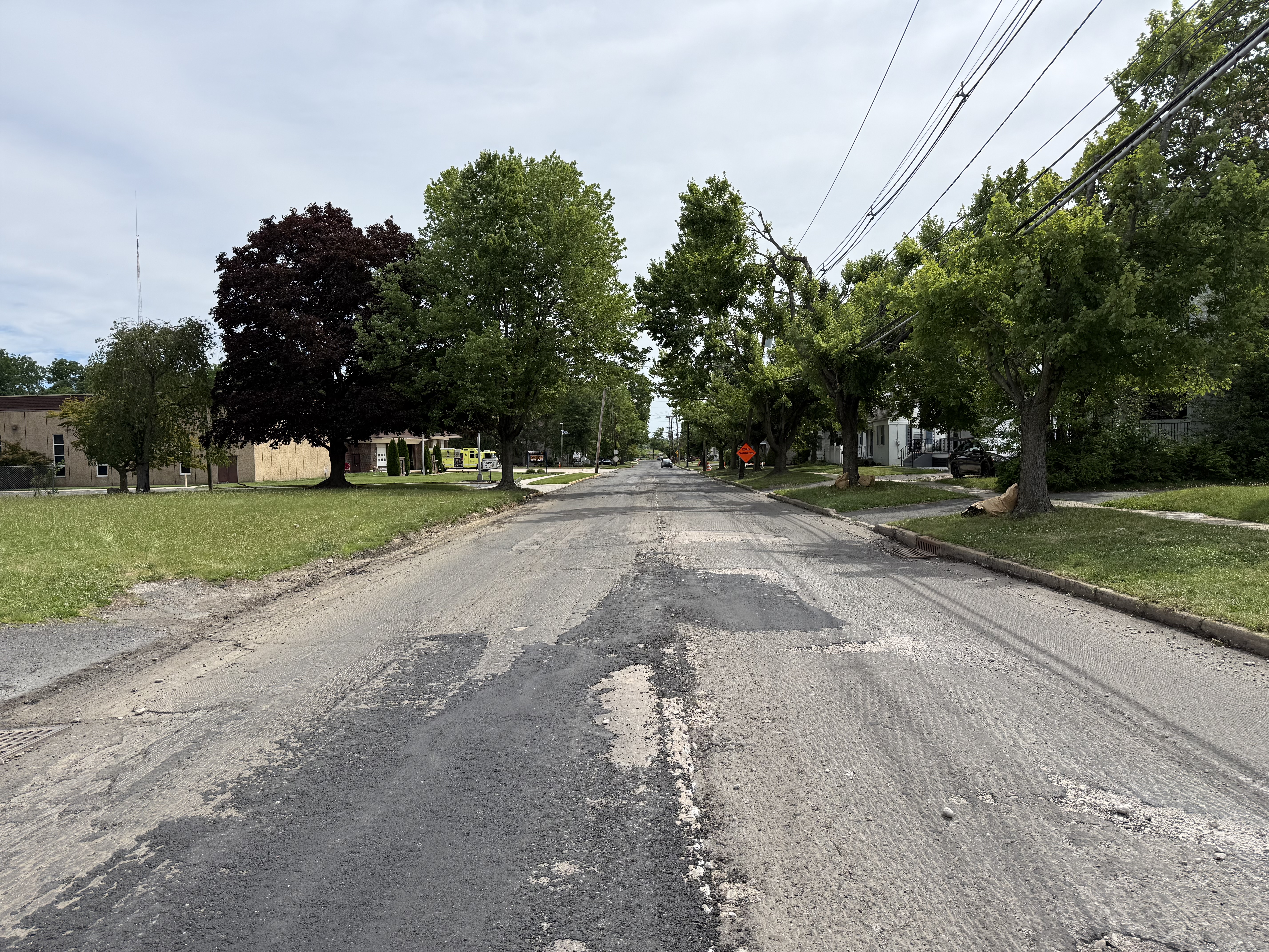 A milled residential street with exposed base material, lined with trees and homes.