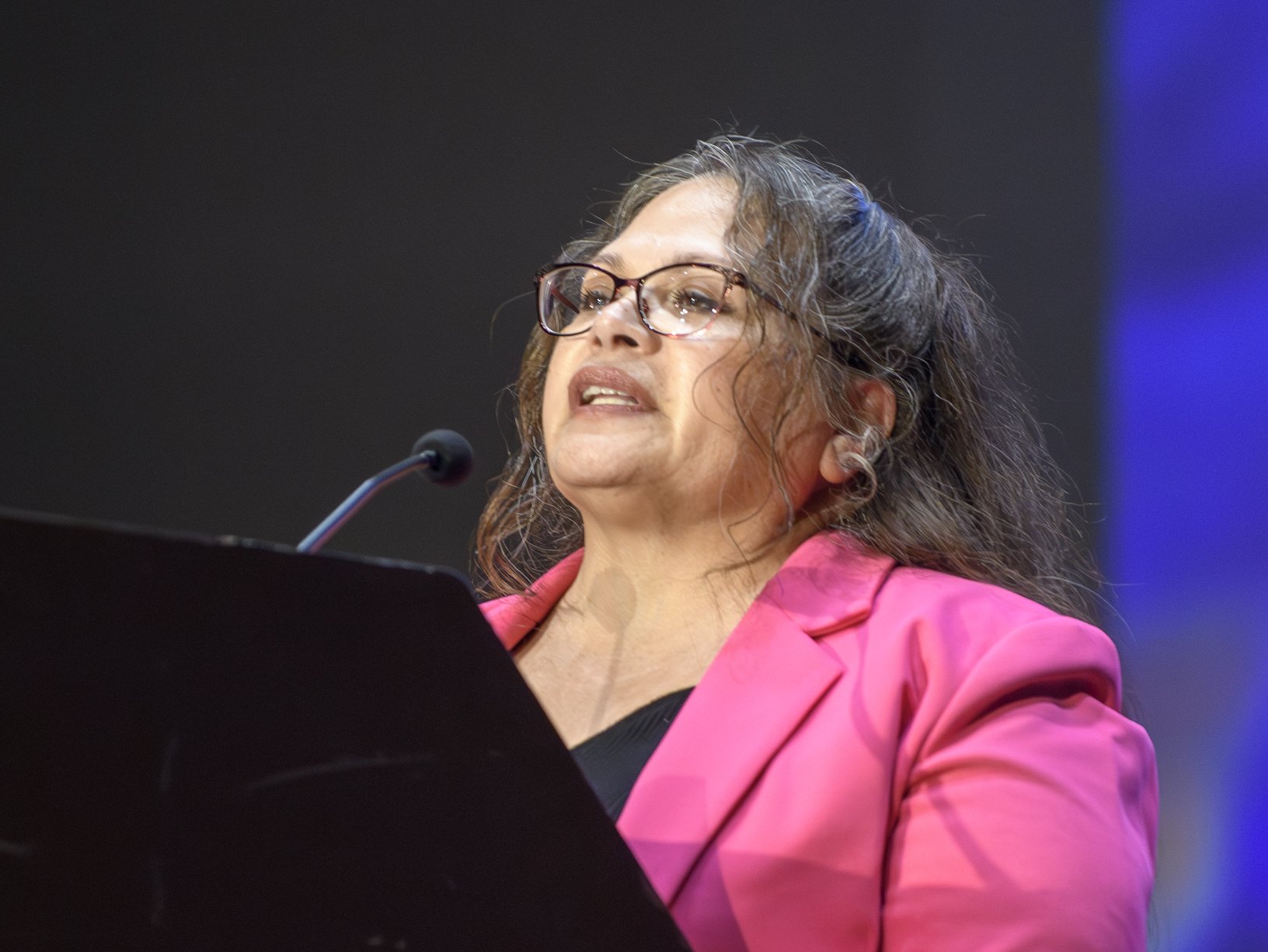 A woman in a pink blazer and glasses speaks at a podium with a microphone.