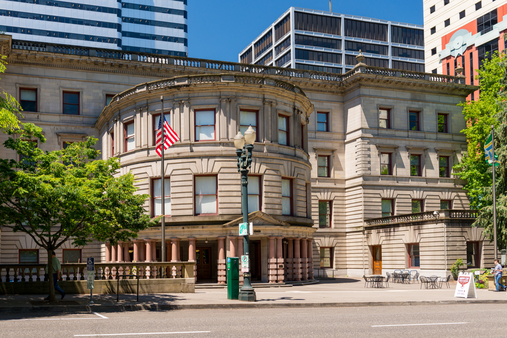 Neoclassical stone government building with American flag and modern towers behind it.