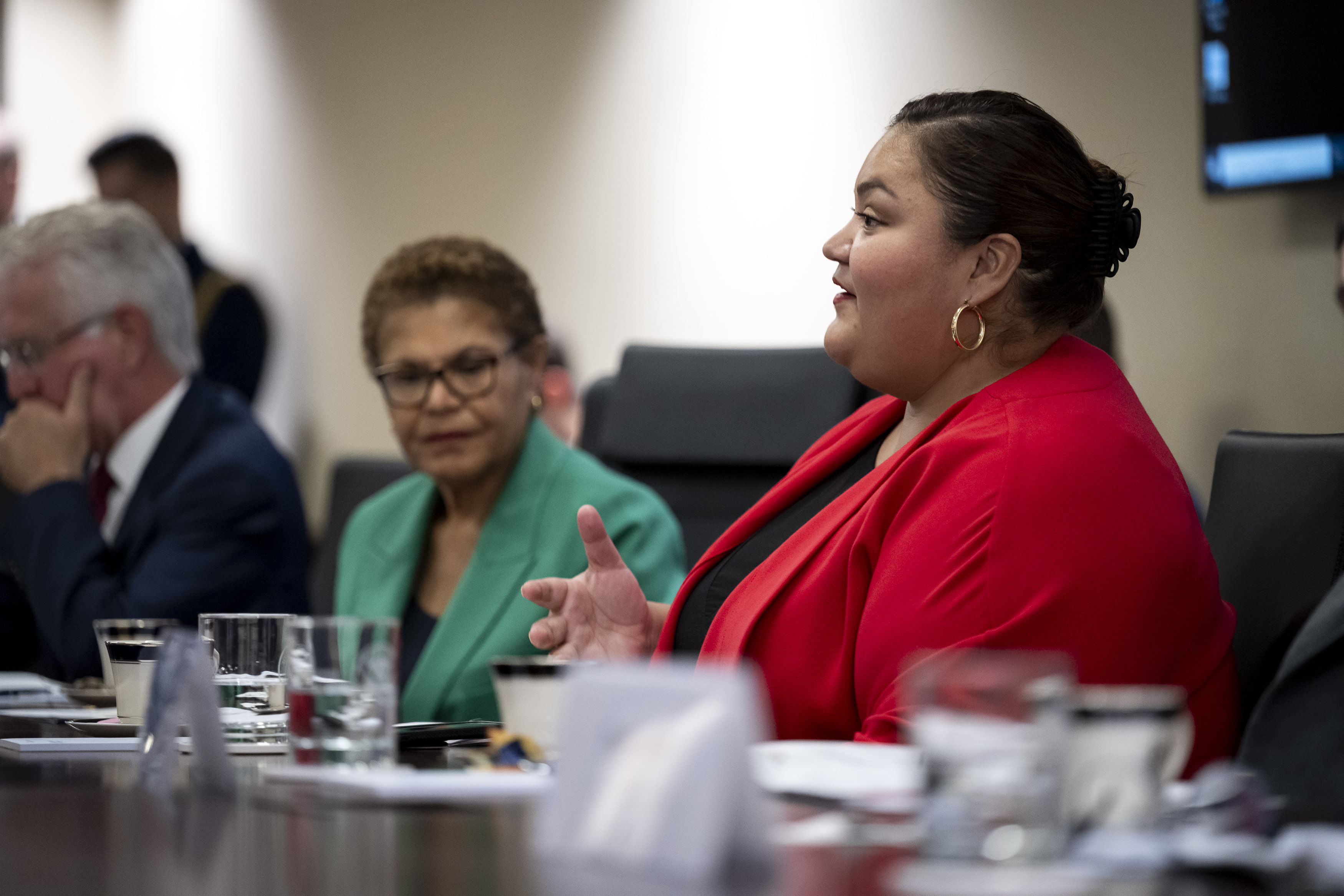 Woman in a red blazer speaking at a conference table during a formal government meeting.