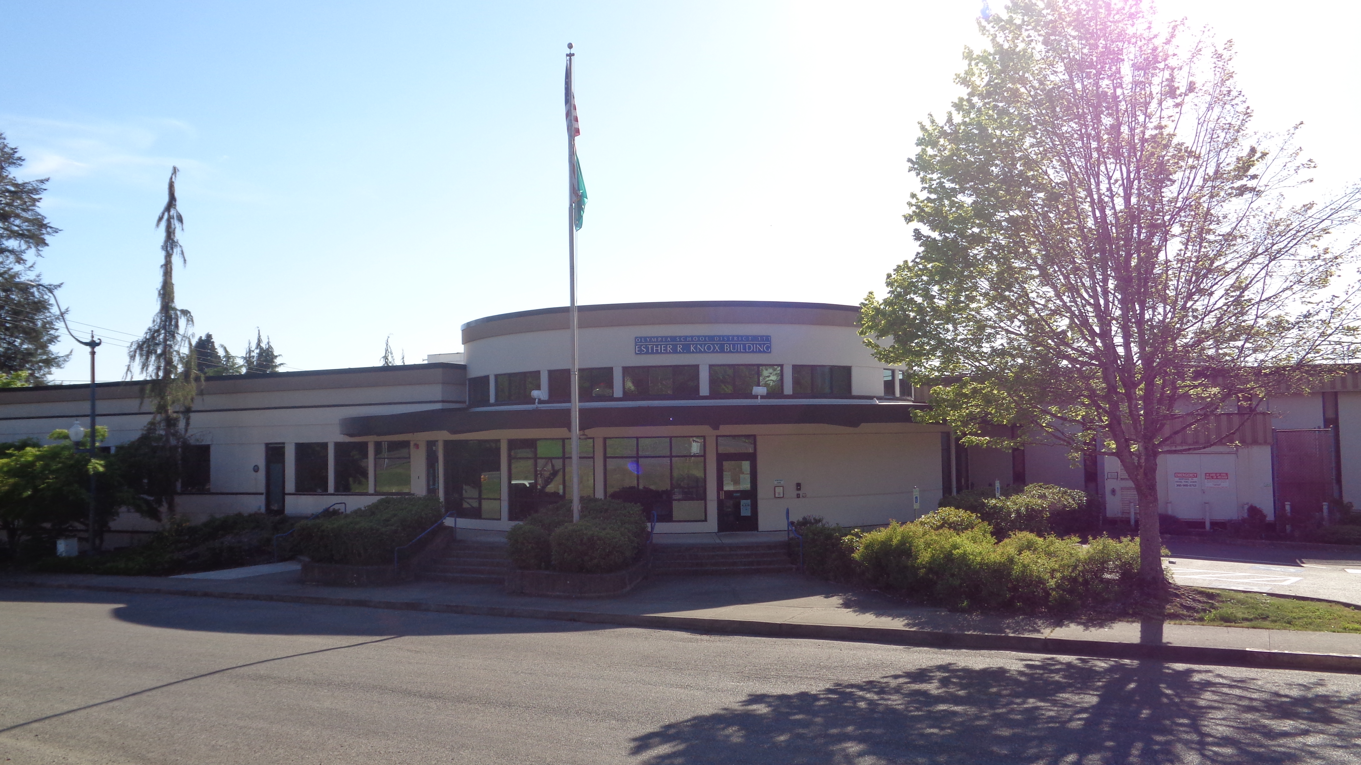 Single-story school district building with curved entrance, flagpole, and trees.
