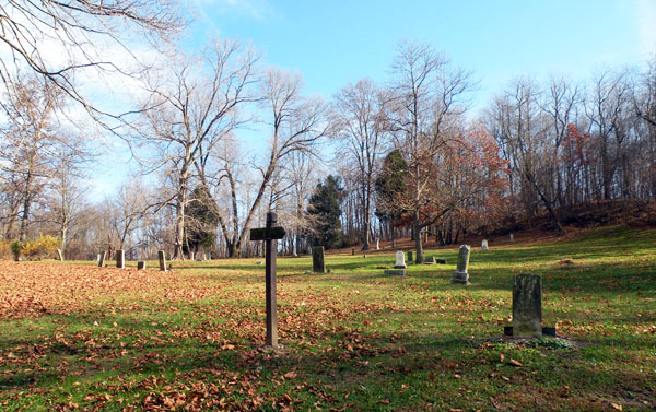 Weathered gravestones and a wooden cross marker in an old cemetery with bare trees.