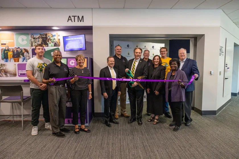Group of people cutting a purple ribbon in front of an ATM alcove indoors.