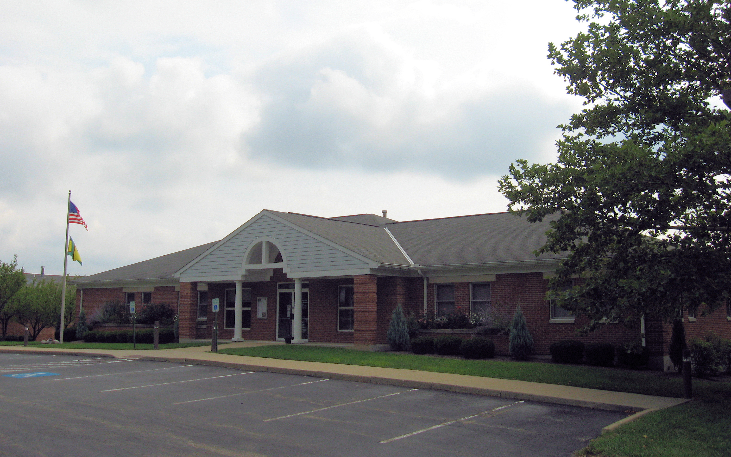 Single-story brick government building with flags on a pole and a parking lot in front.