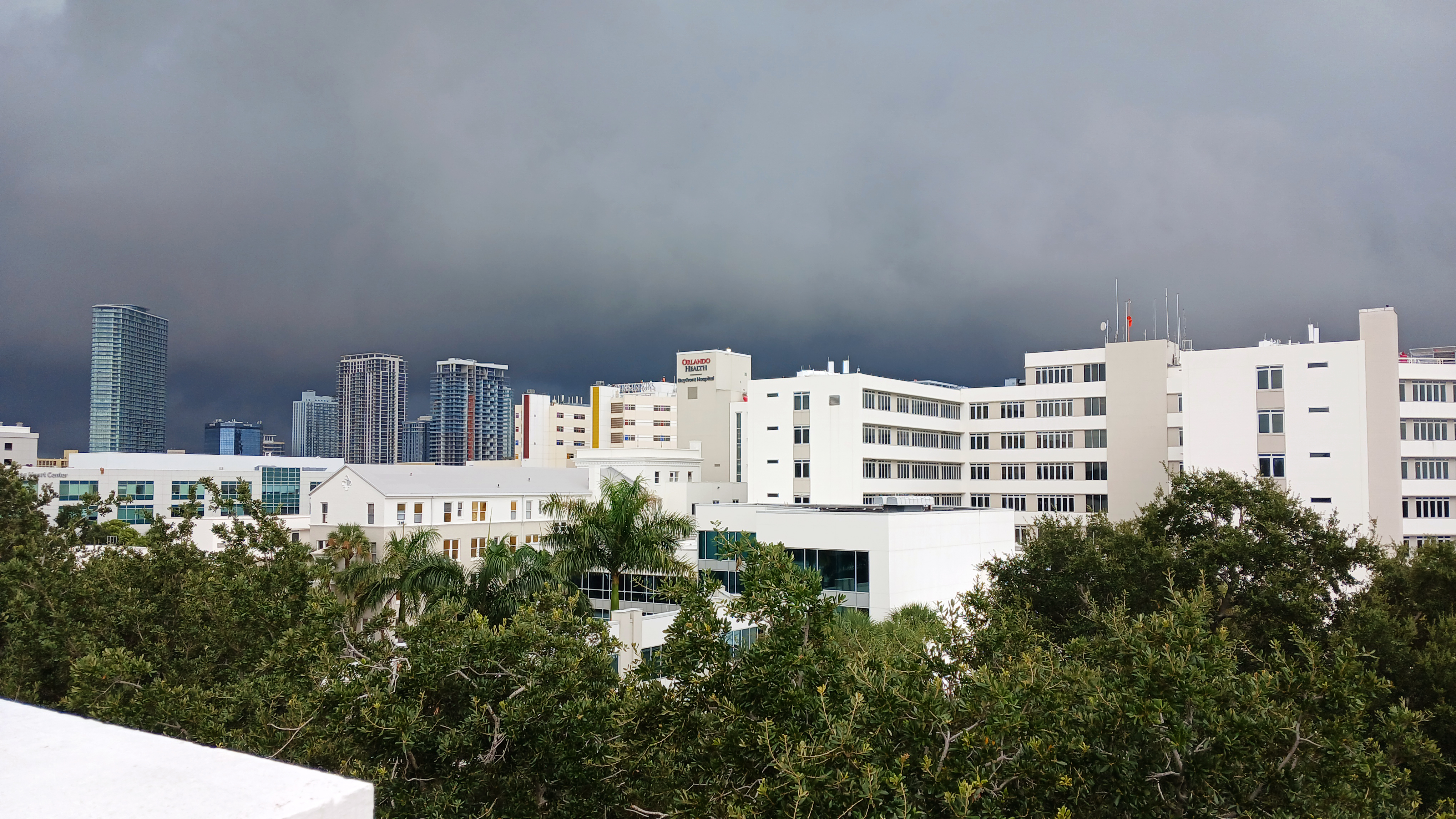 Large white multi-story hospital complex under dark storm clouds, viewed from a rooftop.