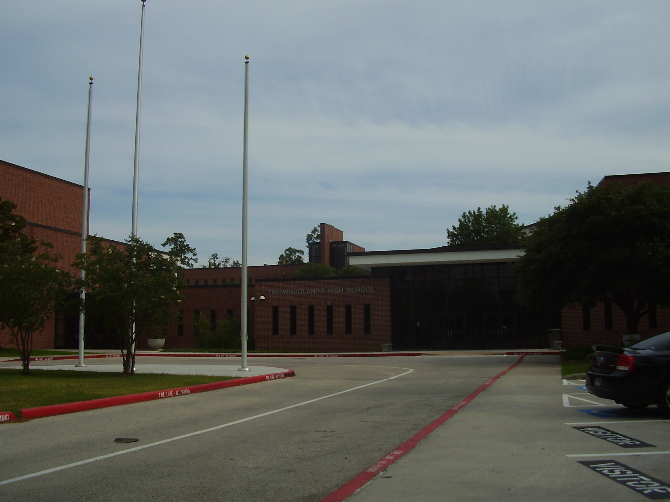 Exterior of a large red-brick school building with three flagpoles in a parking lot.