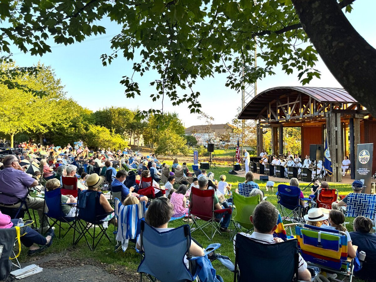 A large crowd seated in colorful lawn chairs faces a covered wooden outdoor stage surrounded by trees.