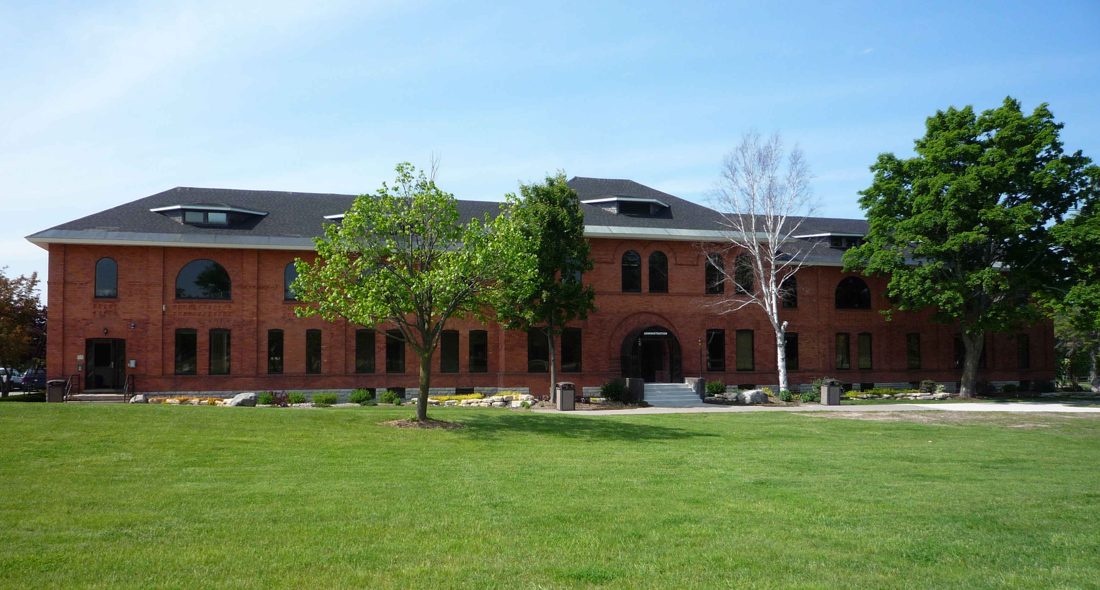 Two-story red brick building on a university campus with green lawn and trees under blue sky.