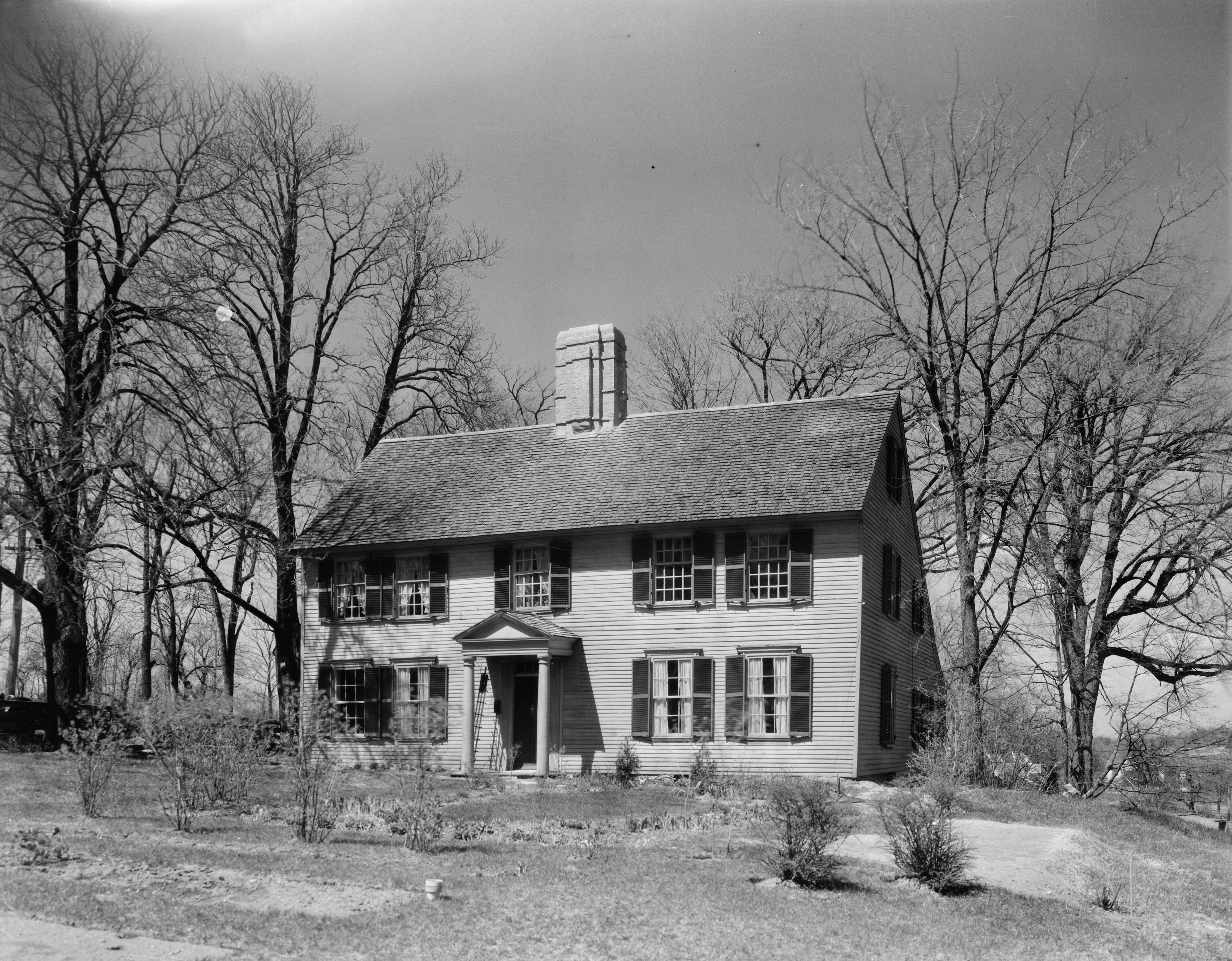Two-story colonial wood-frame house with shuttered windows and central chimney, photographed in black and white.