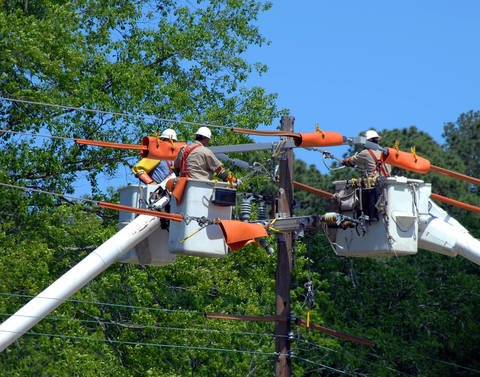 Two utility workers in bucket trucks repairing power lines near a wooden pole surrounded by trees.
