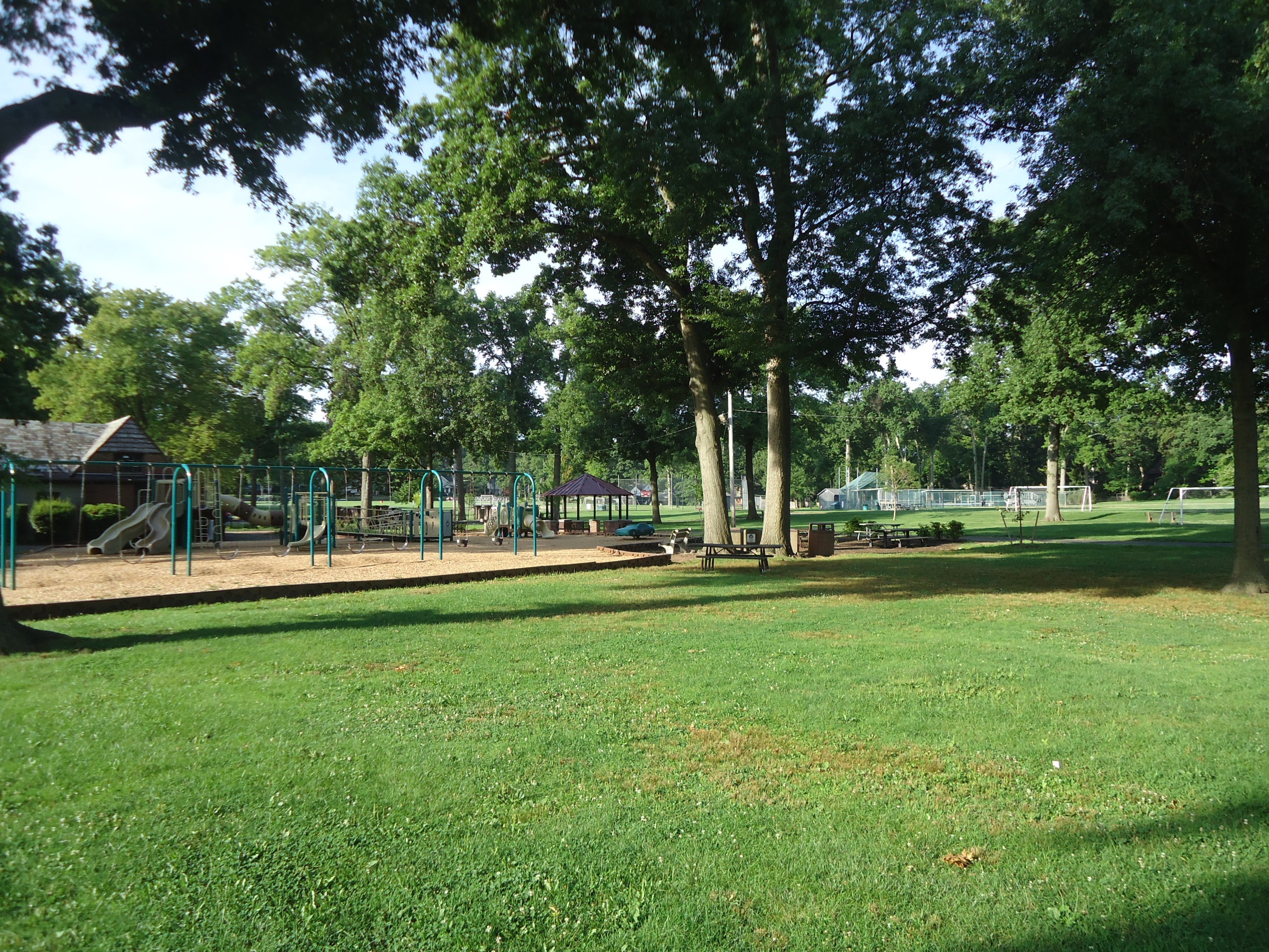 A playground with climbing structures on mulch surrounded by mature trees and a green lawn.