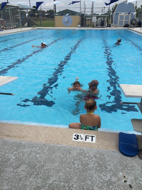 Children and an adult at a pool during swim lessons, with one child sitting on the pool edge.