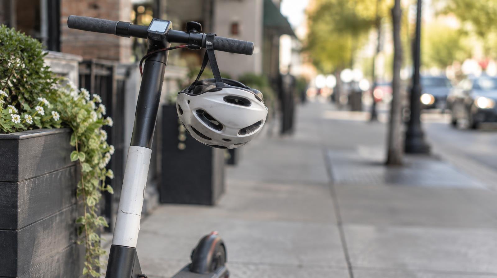 An electric scooter parked on a sidewalk with a bicycle helmet on the handlebars.