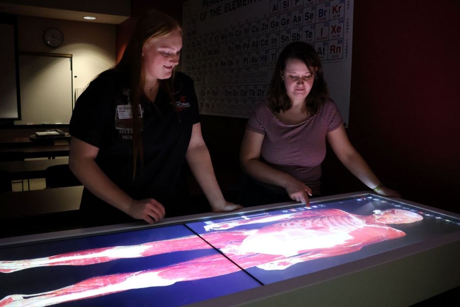 Two people lean over a large glowing touchscreen displaying a human anatomical diagram.