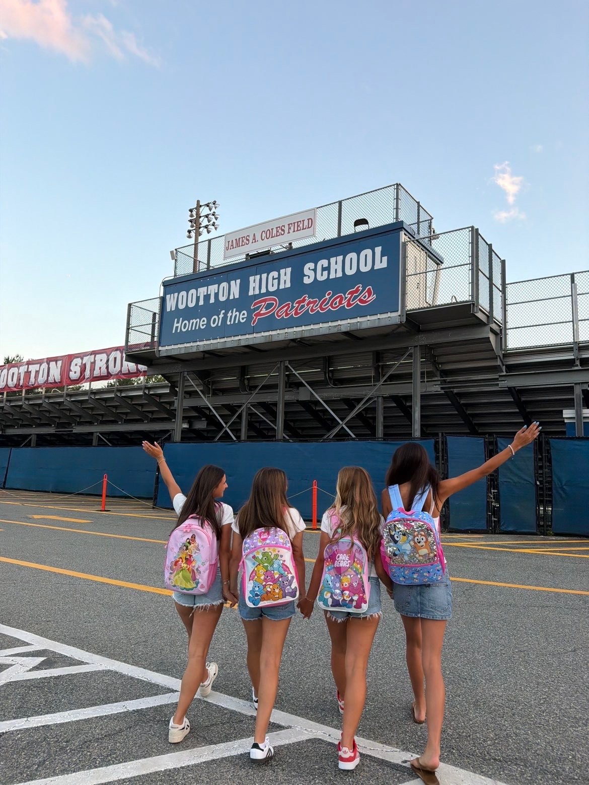 Four students seen from behind wearing colorful cartoon backpacks in front of a stadium sign.