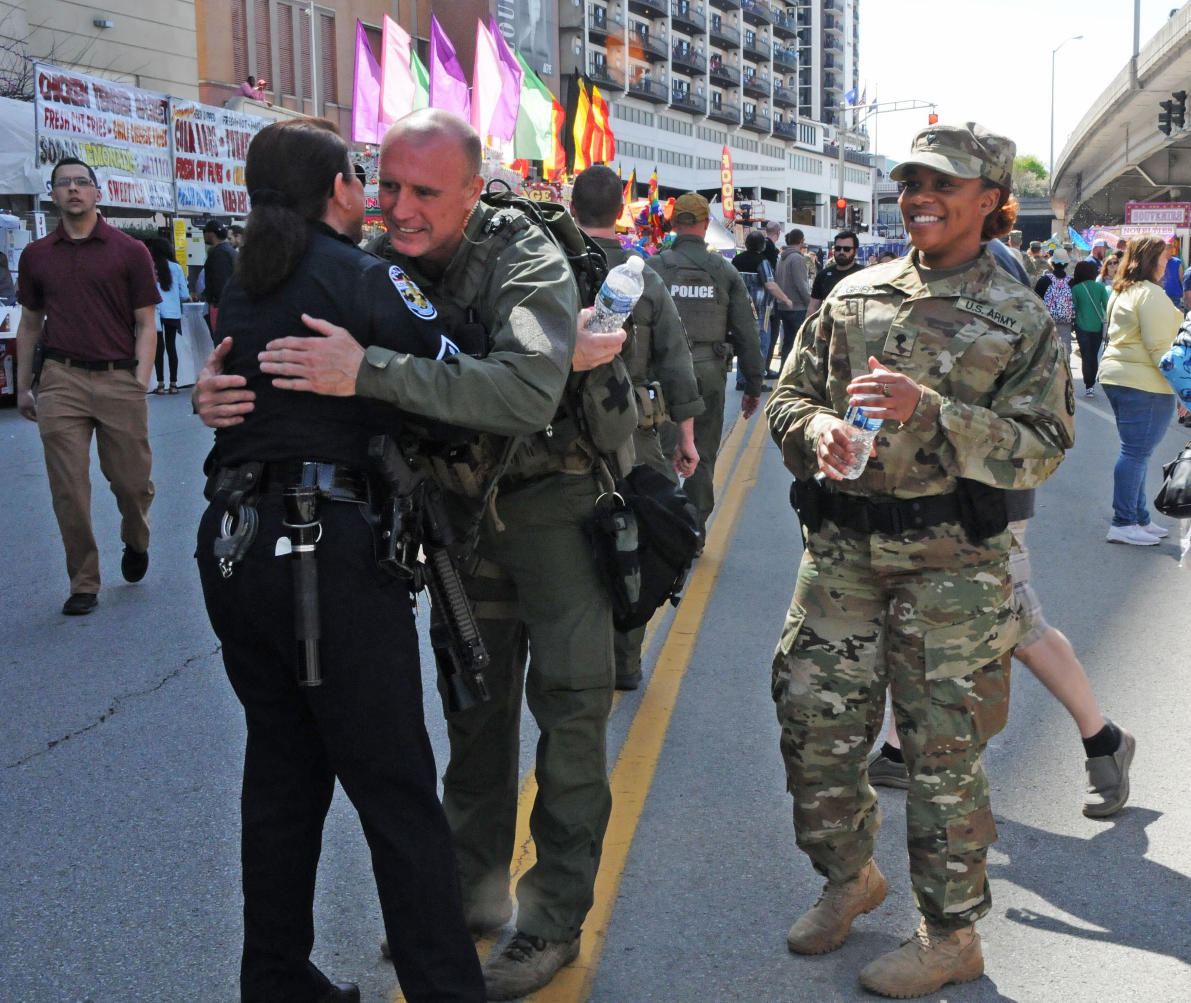 A police officer and a SWAT officer embrace on a city street while a soldier in camouflage smiles nearby.