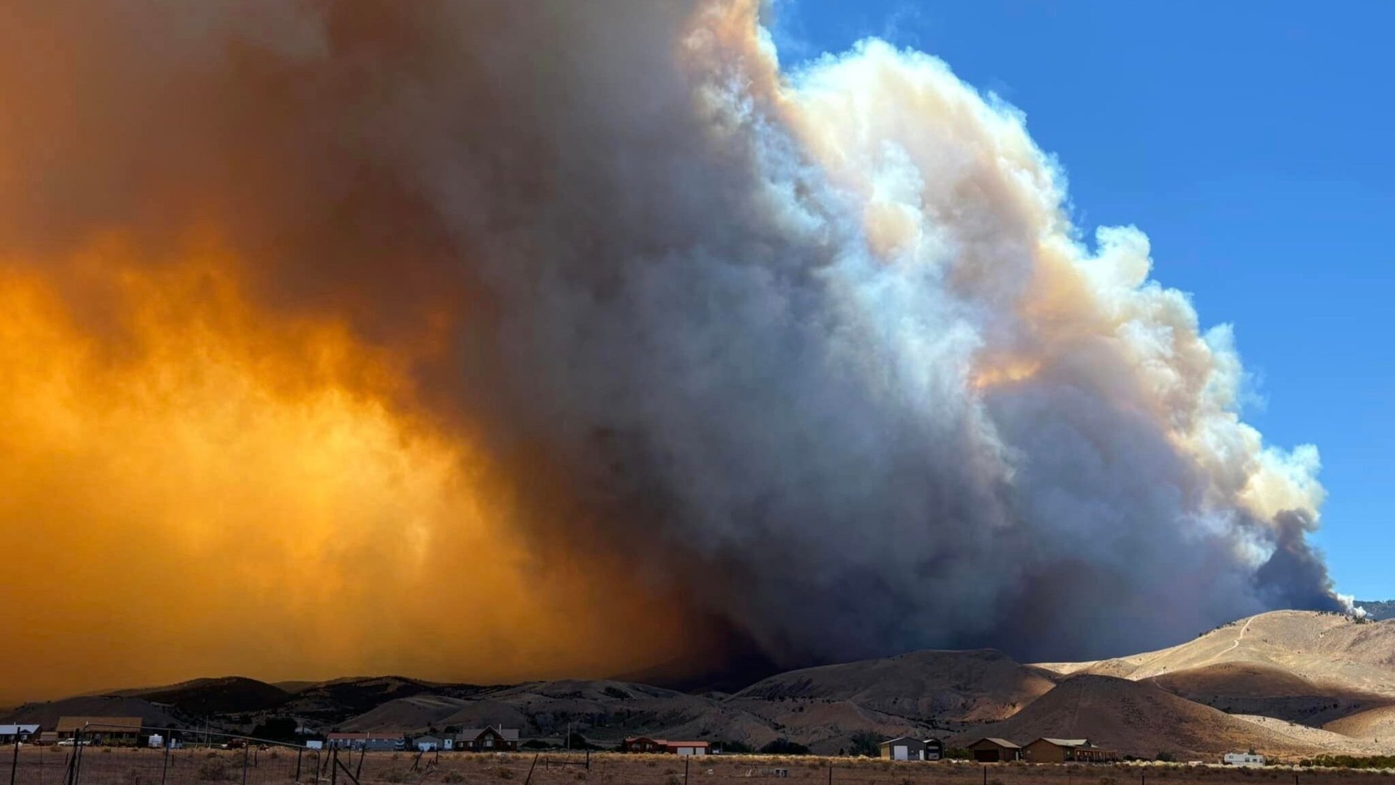 Large orange and gray smoke plume rises from a wildfire over dry desert hills.