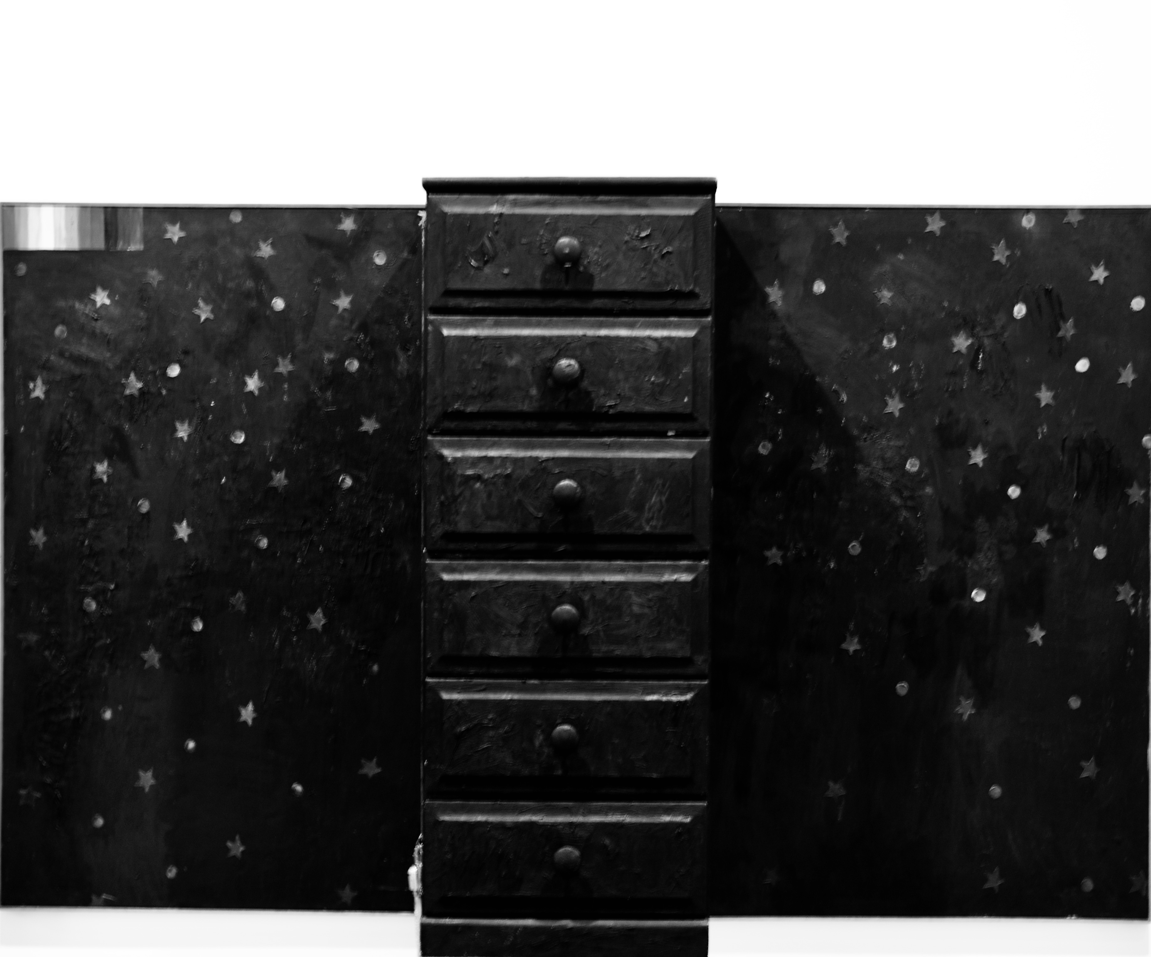 A wooden chest of drawers mounted on a dark star-patterned canvas in a gallery.