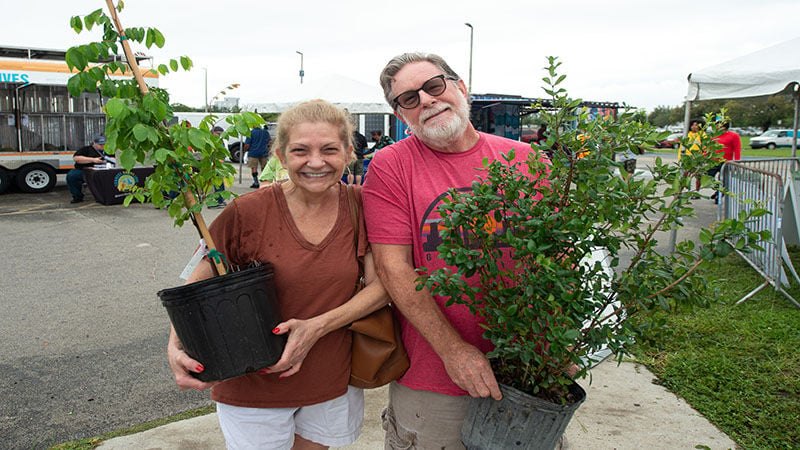 Two smiling people hold small potted trees at an outdoor community event.