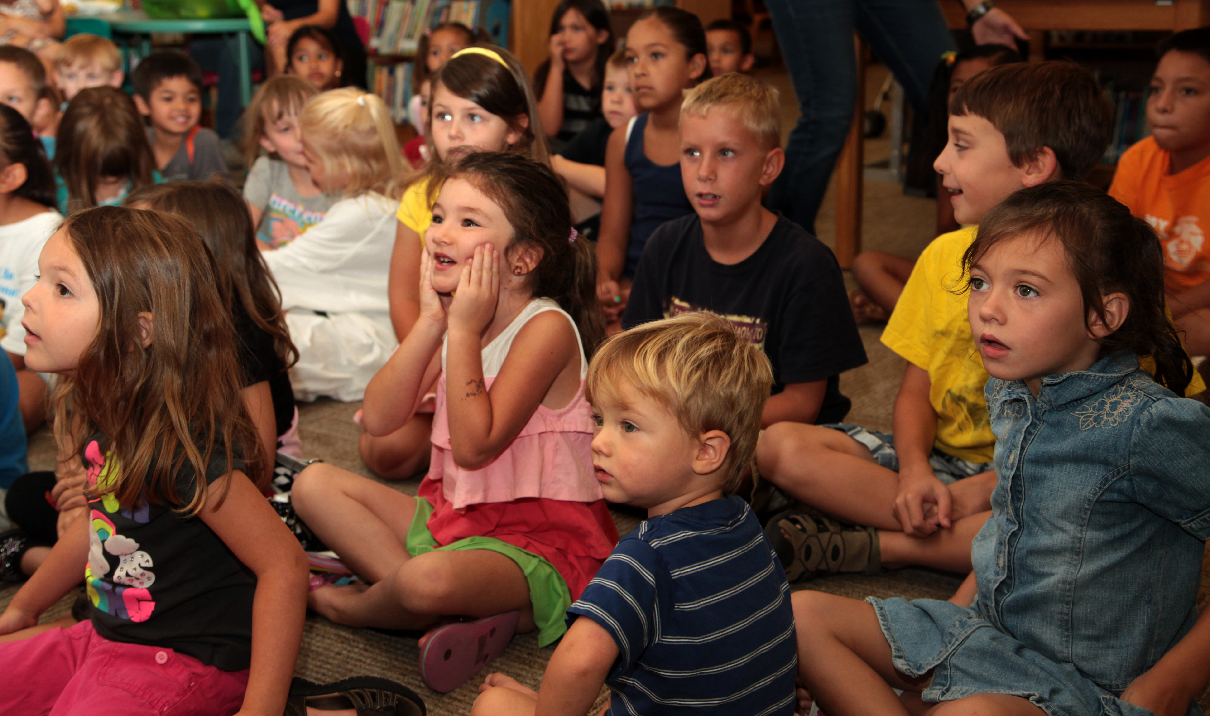 A group of young children sit on a floor with excited, wide-eyed expressions during a library program.