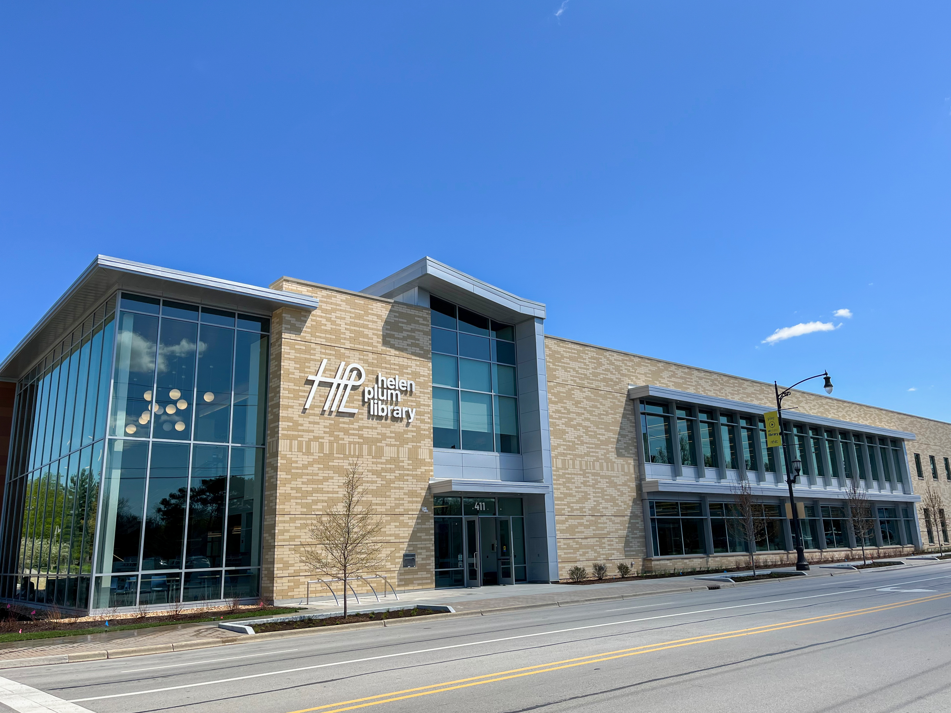 Modern two-story library building with tan brick and large glass windows on a street corner.
