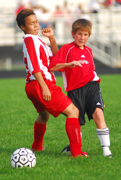 Two young male soccer players in red uniforms compete for a ball on a grass field.