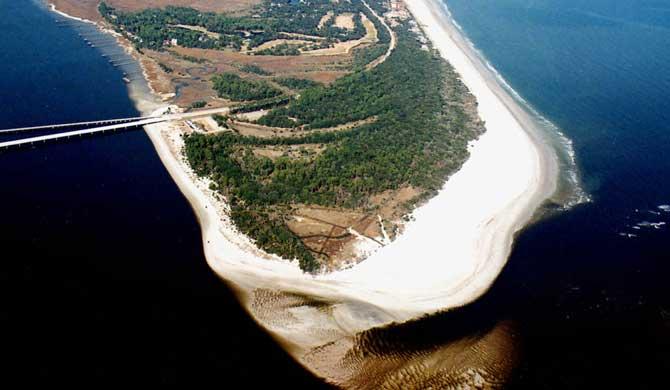 Aerial view of a narrow barrier island with white sand beaches and dense tree cover.