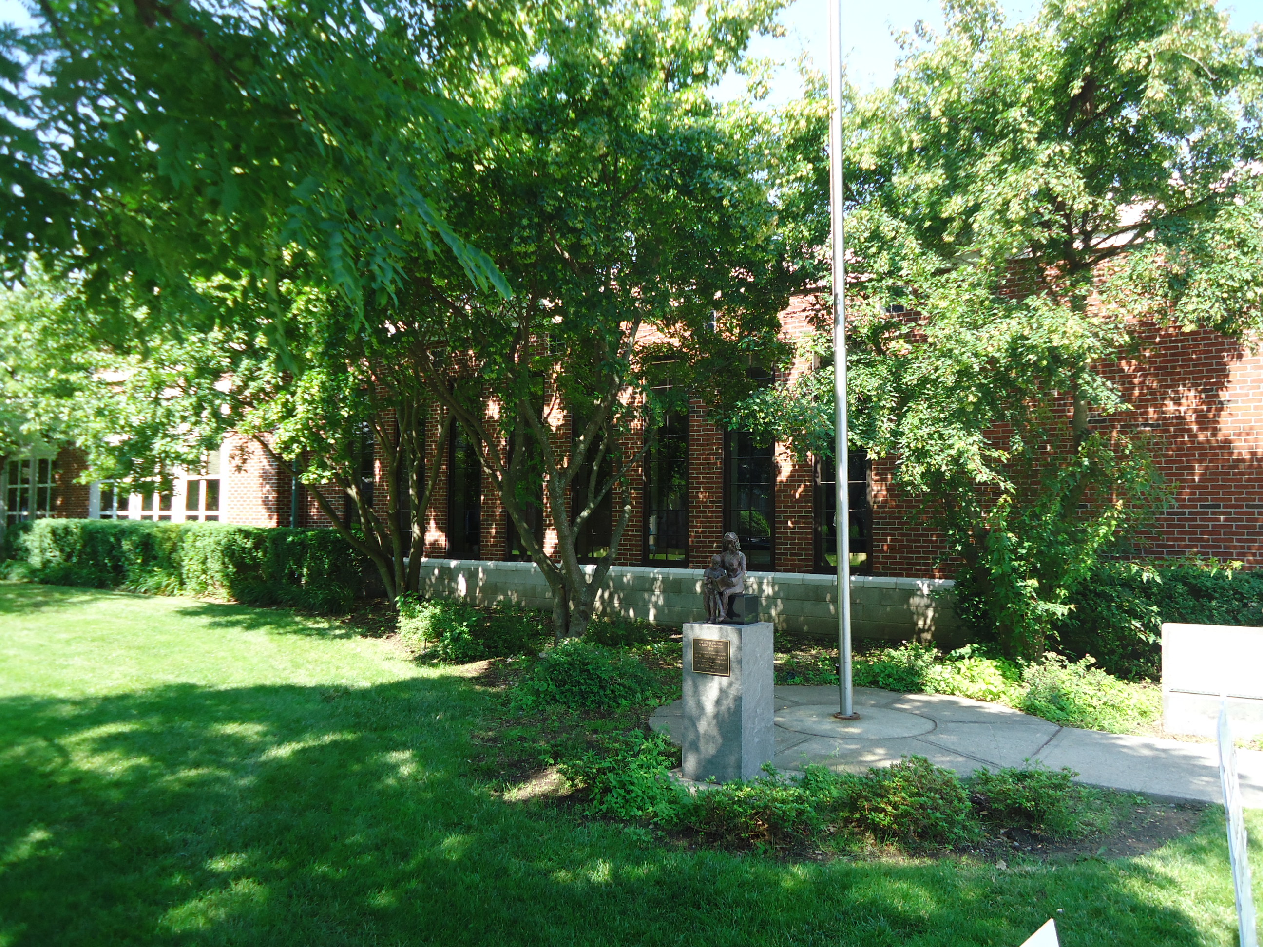 Brick public library building partially visible behind a canopy of mature trees.