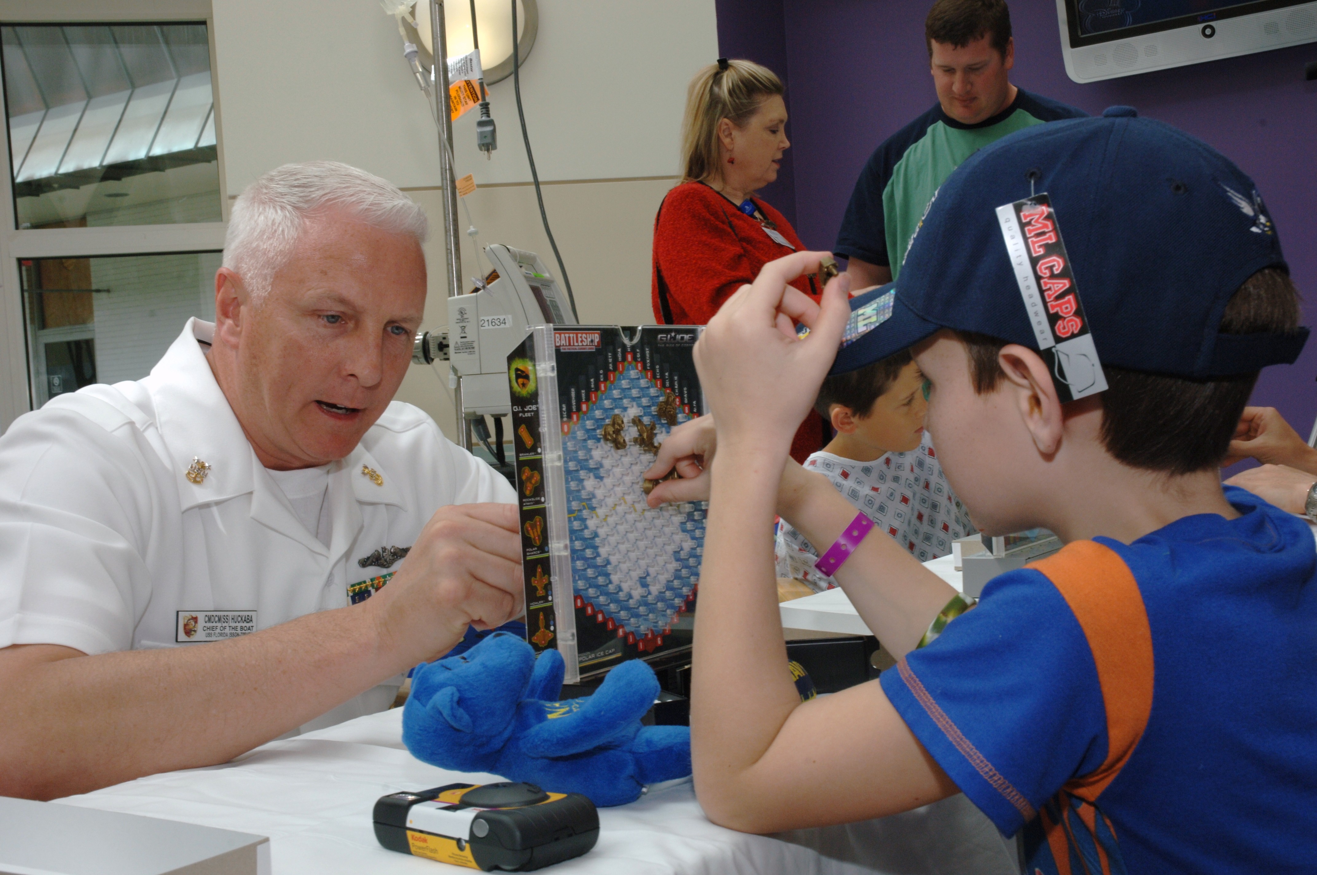A man in a white uniform plays a board game with a young child at a hospital table.