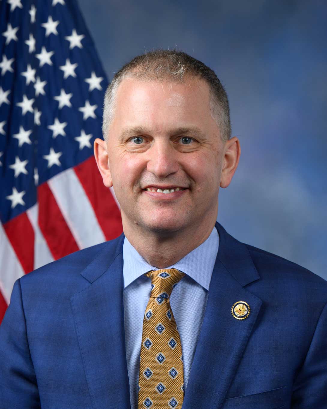 Man in blue suit and gold tie smiling before an American flag.