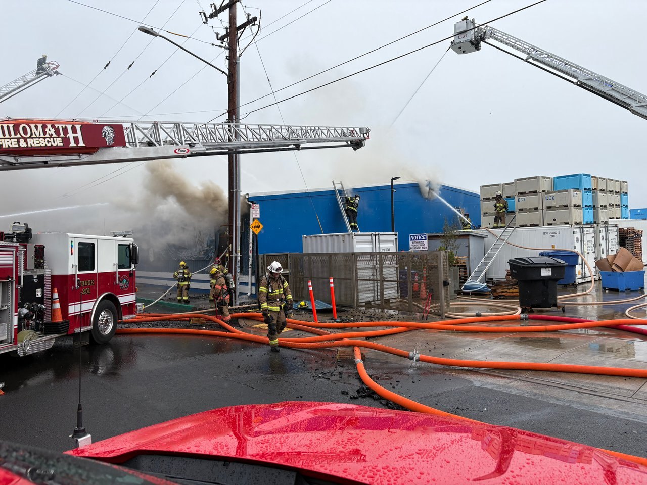 Firefighters in turnout gear work at a large industrial building fire with aerial ladders extended and smoke billowing.