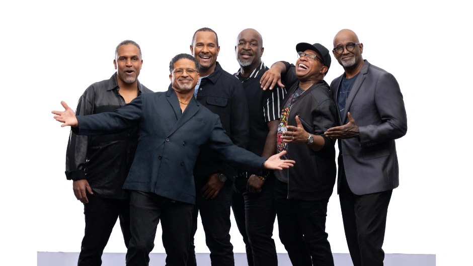 Six men in casual and dark attire pose together smiling against a white background.