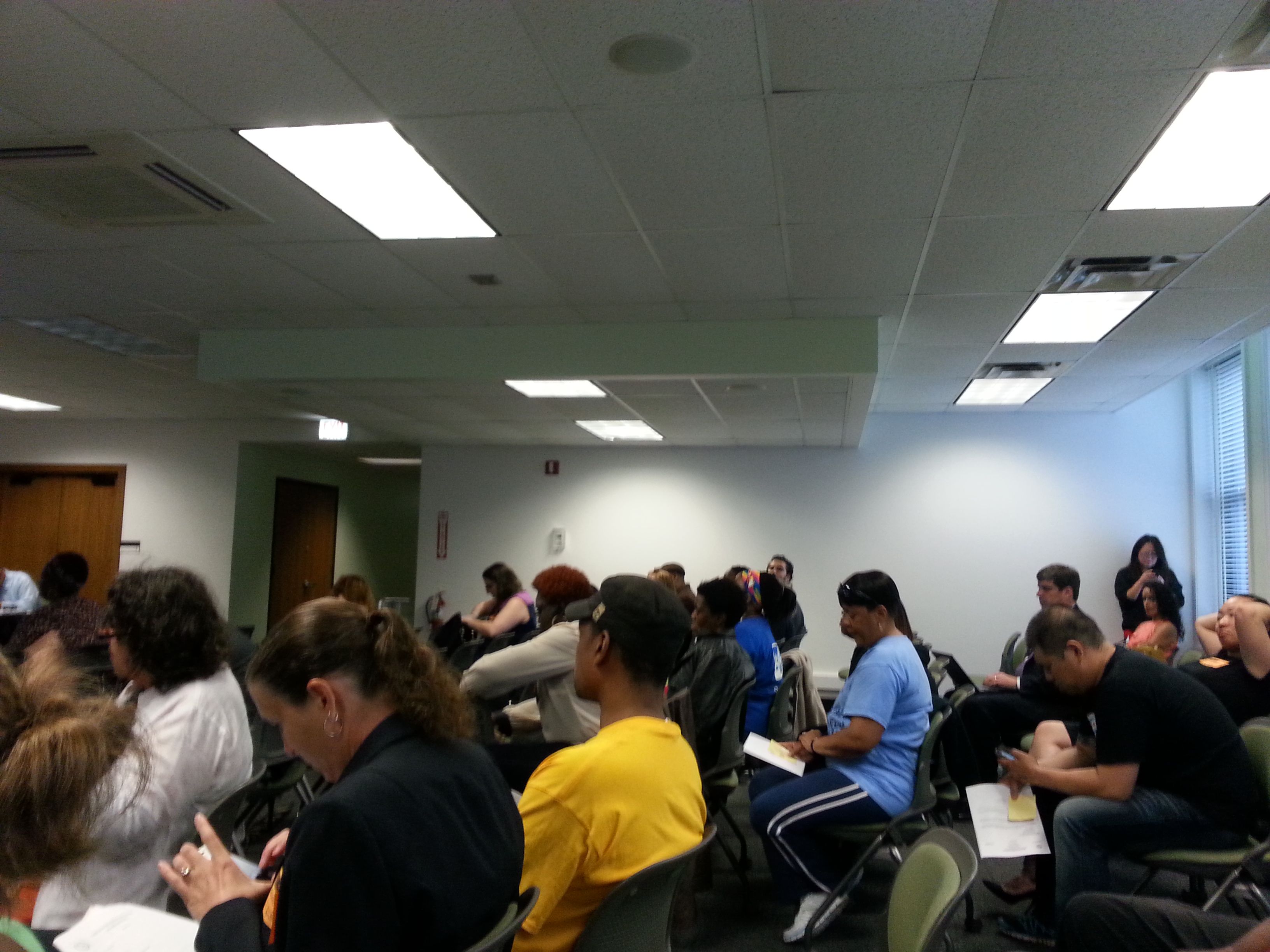 Crowded meeting room with rows of people seated in chairs and one person standing at the front.
