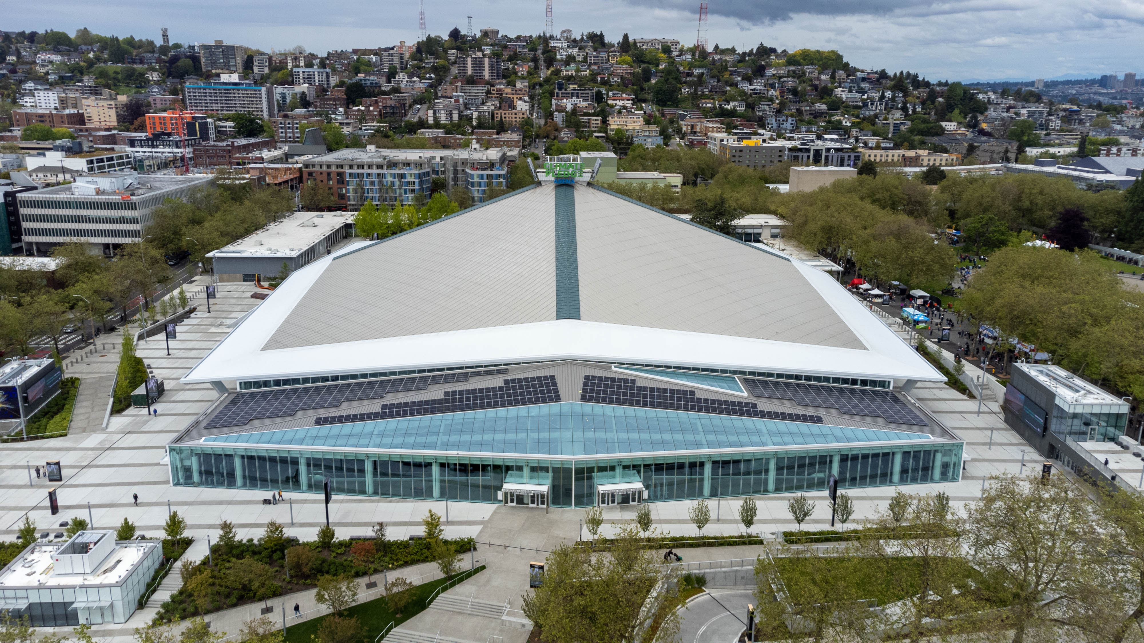 Aerial view of a large arena with a white peaked roof and surrounding plaza.