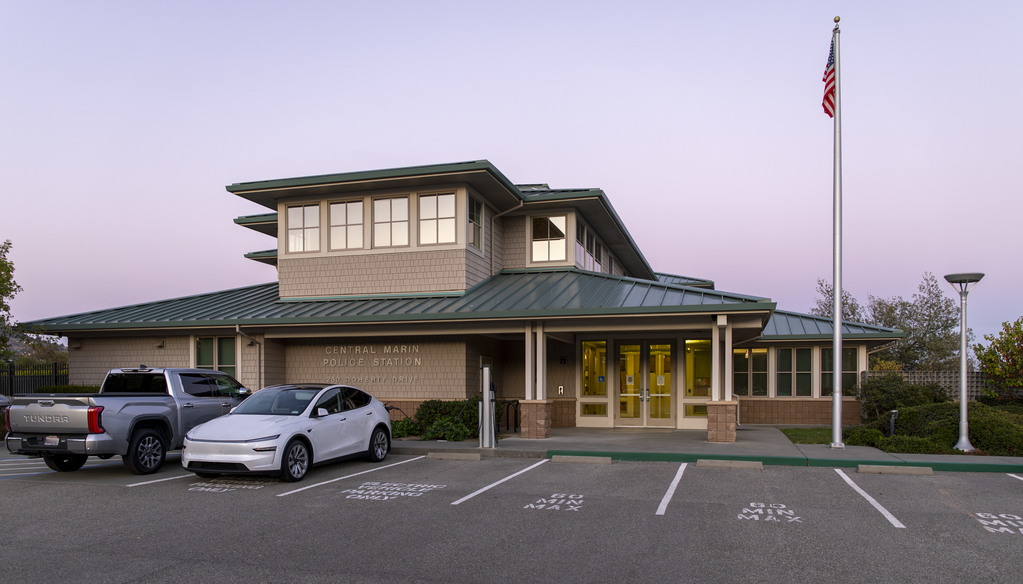 Two-story government building with green metal roof and American flag at dusk.