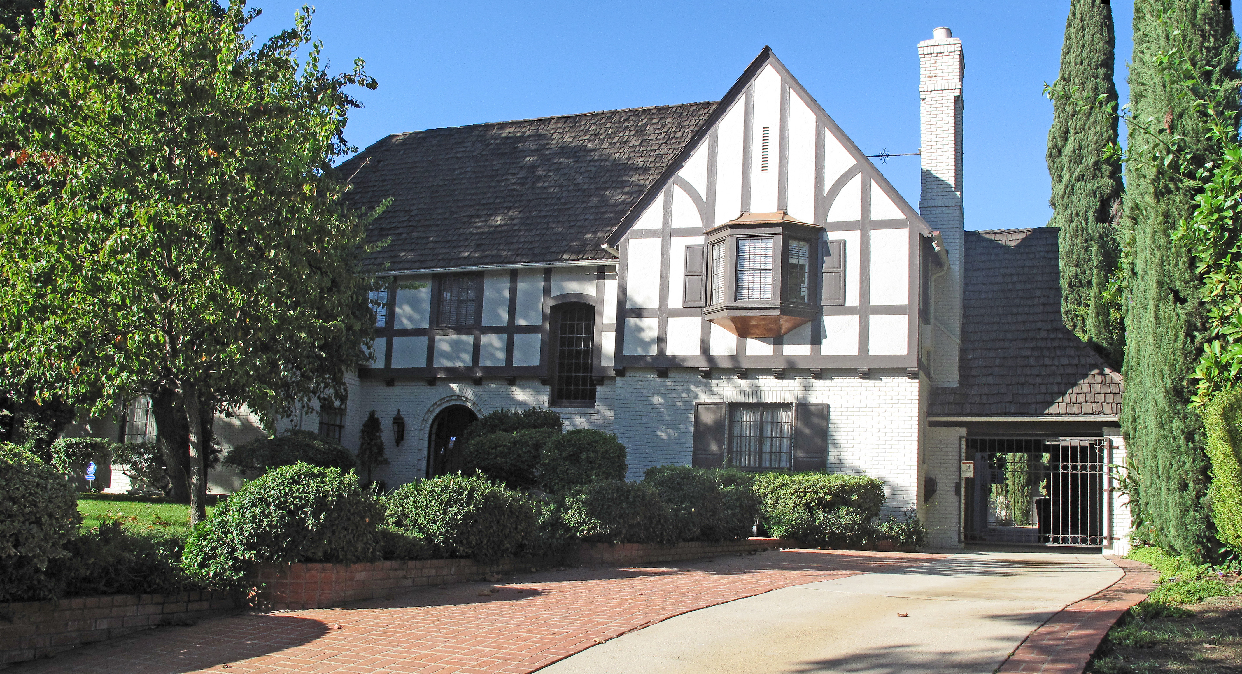 Two-story Tudor Revival house with white stucco, dark timber framing, and cypress trees.