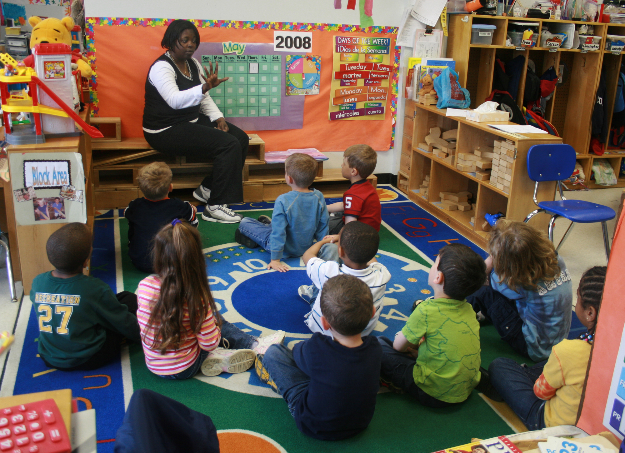 A teacher sits on steps addressing young children seated on a colorful rug in a classroom.