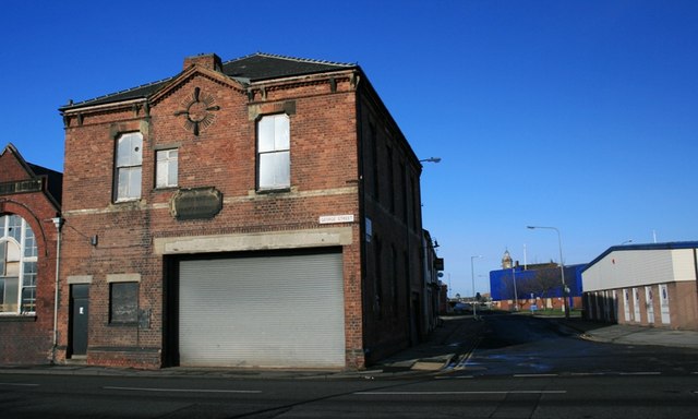 A derelict red-brick fire station building on a street corner with a roller-shutter garage door and boarded windows.