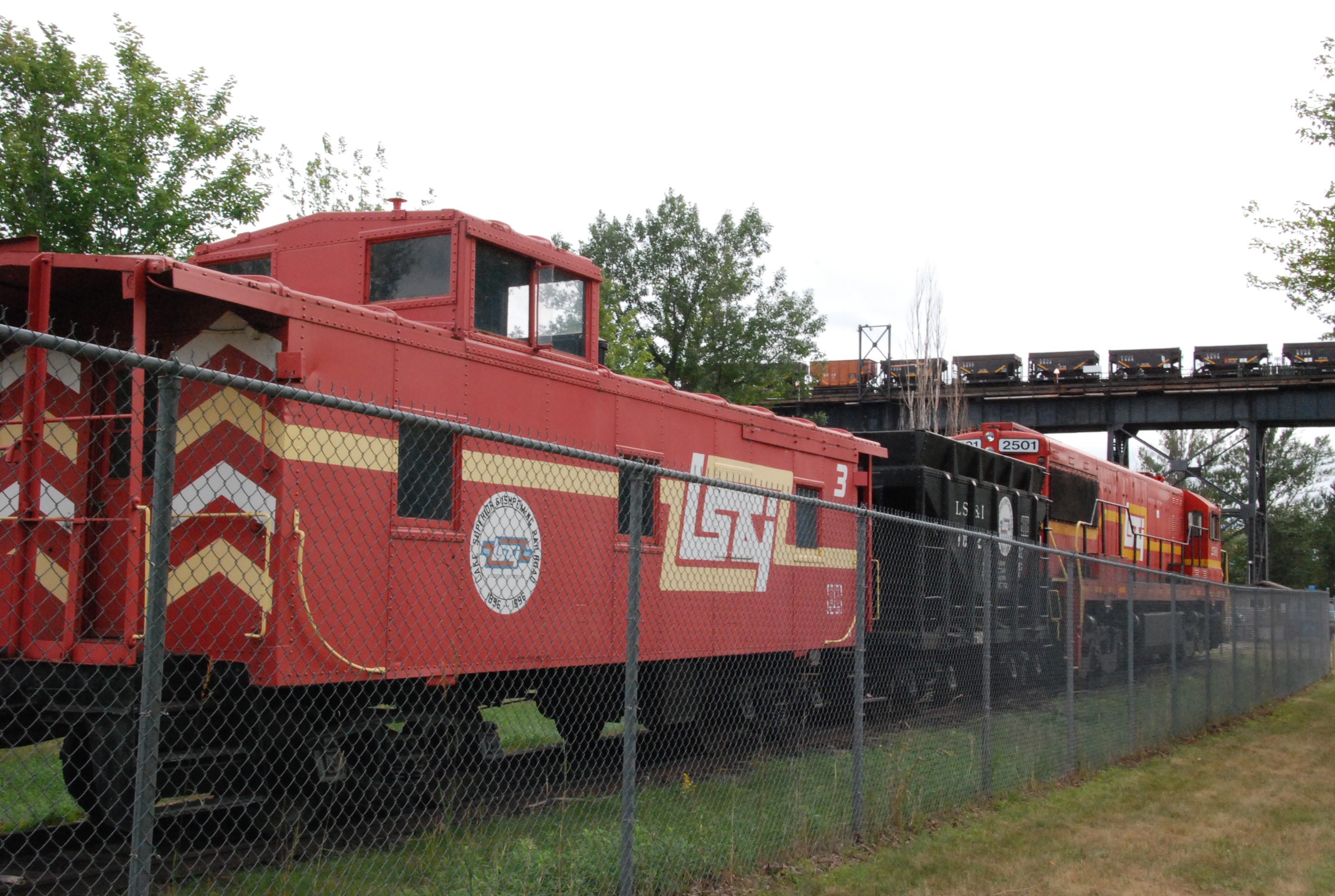 Railroad locomotive and ore hopper cars at an industrial ore dock facility behind a fence.