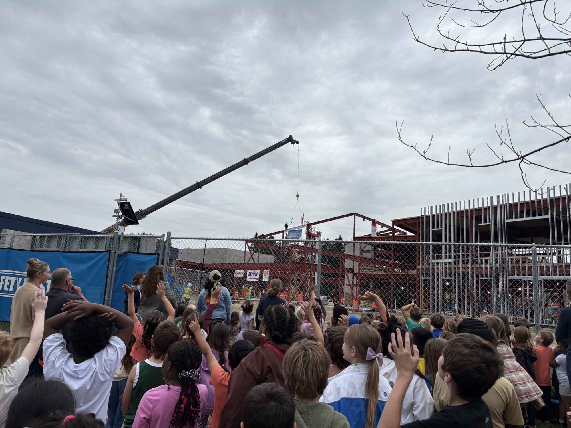 Crowd of children and adults with raised hands watching a crane at a construction site.